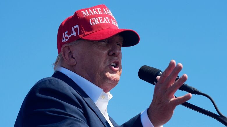 A person wearing a red hat with a slogan on it reading, 'Make America Great Again,' gestures while speaking into a microphone.