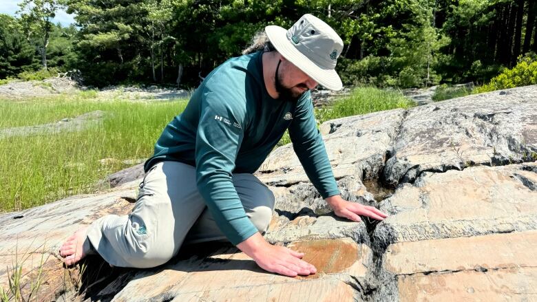  A man in a green long sleeved shirt with a bucket hat sits on a rock and wets down a an area on the rock.