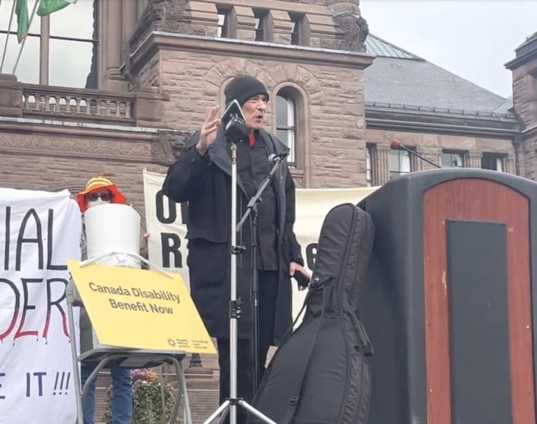 A person speaks at a demonstration. A sign next time to him reads 