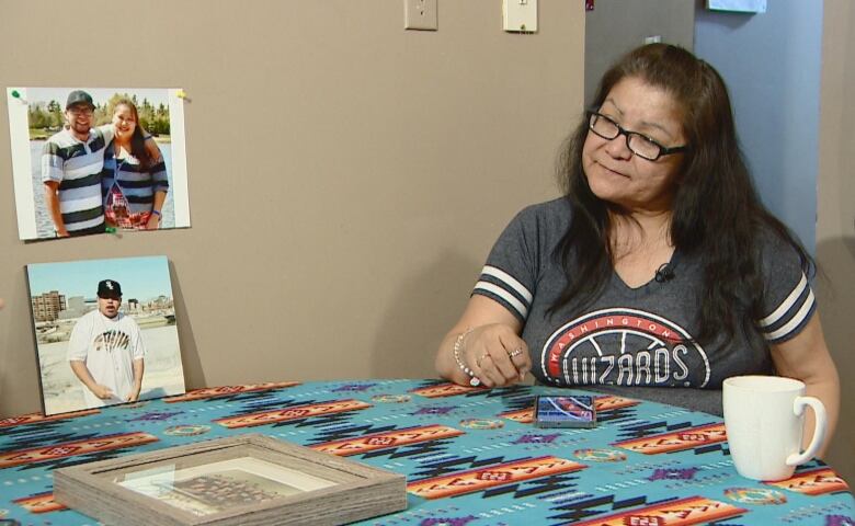 A woman in a dark grey shirt sits at a table, with two pictures hung up beside her, one showing a couple and another showing a man in a black cap.