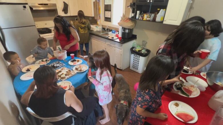 A woman in a red shirt can be seen in a kitchen, with multiple children seen all around her.