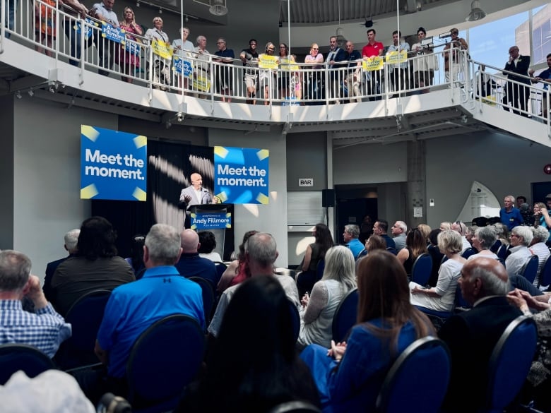 A man wearing a suit stands before a podium and speaks to a crowd of people gathered in front of him and on a balcony above him.