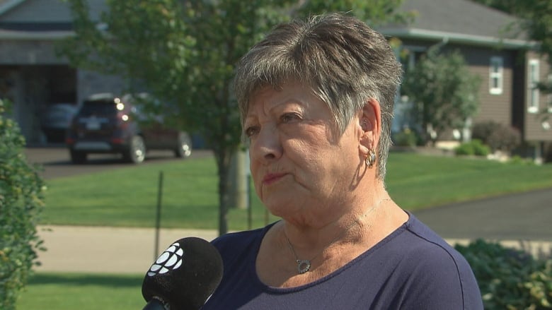 A woman with short hair speaks into a microphone in a suburban community of homes.