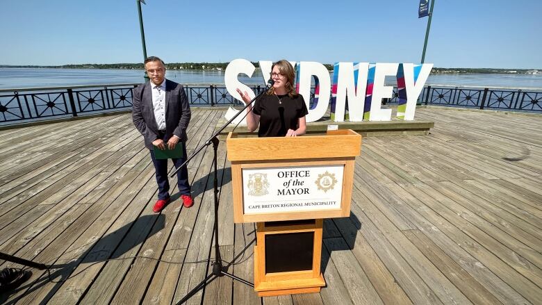 A man with a dark blue suit, red shoes and black glasses stands off to the side as a woman in a black shirt and glasses speaks at a podium on a wooden deck overlooking a harbour.