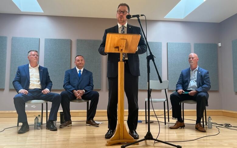 A man in a business suit stands speaking at a podium as three other men sit on chairs behind him. 