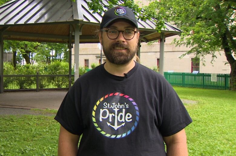 A man wearing a black t-shirt and black hat standing in a park. 