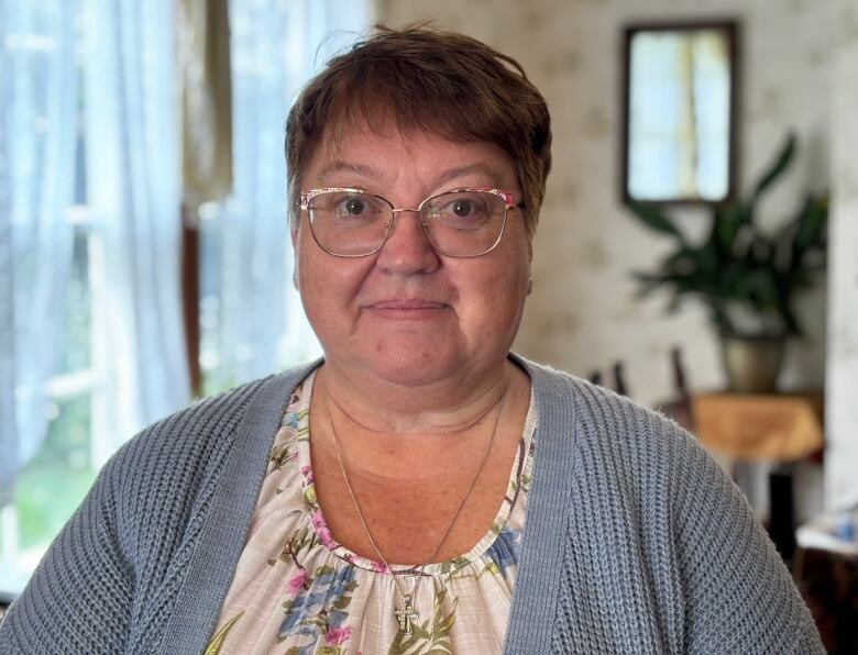 A woman wearing glasses and a blue cardigan stands in a living room.