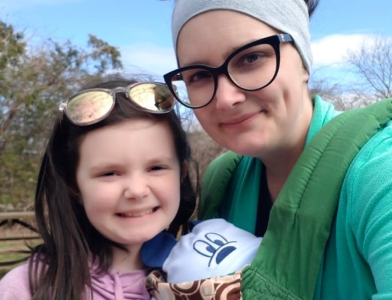 A young girl with brown hair poses for a selfie with her mother. Her mother wears thick black glasses and a grey headband. 