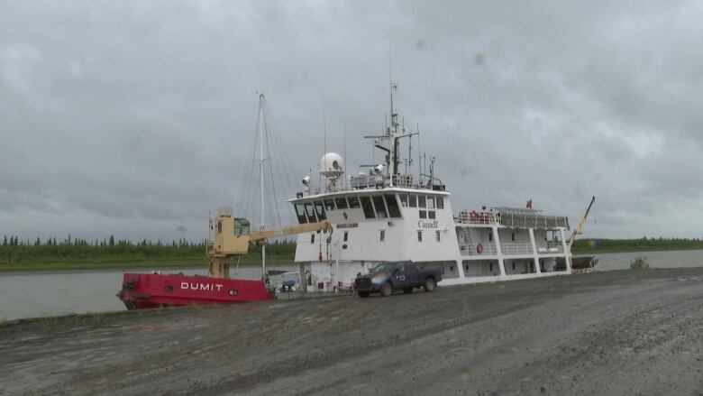 A coast guard boat on a river. It's pulled up next to a gravel road with a black truck. 