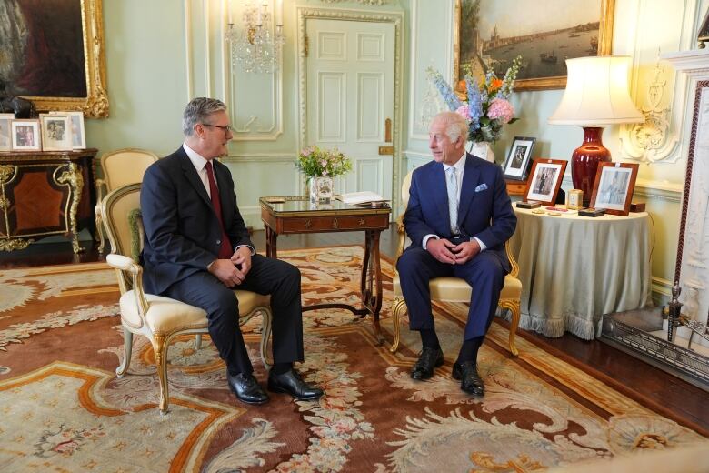 Two men sit next to each other in a carpeted room while speaking.