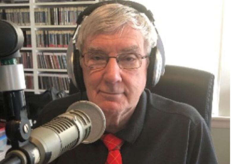 A man with glasses sits behind a microphone in a radio station.