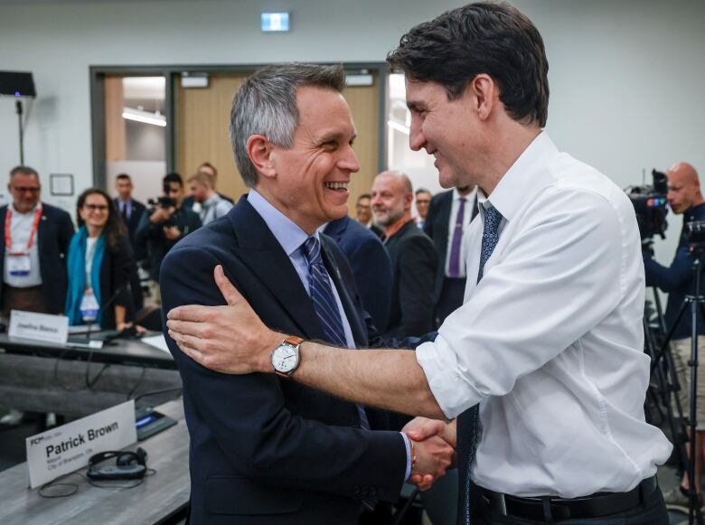 A smiling man with grey hair in a blue suit shakes hands with a dark-haired man in a white shirt with the sleeves rolled up