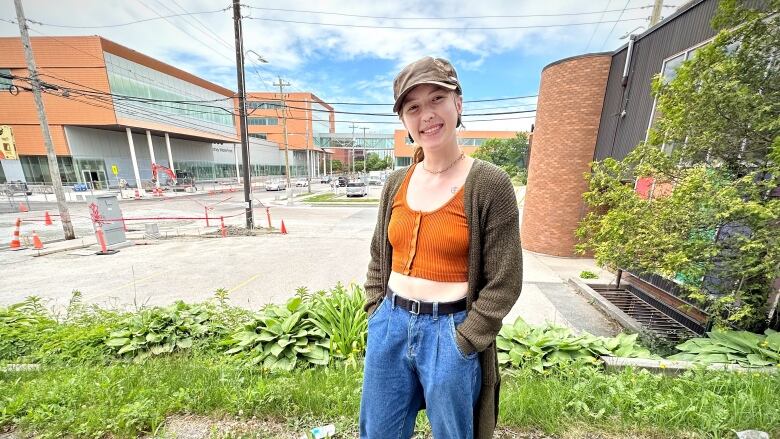 A young woman in blue jeans, an orange tube top and a long brown sweater wearing a ball cap stands in front of some buildings under construction.