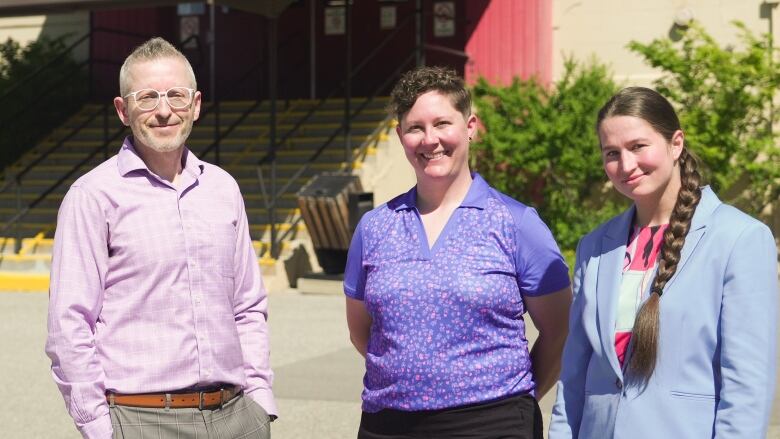 A man wearing a pink shirt, a woman wearing a purple shirt, and a woman wearing a blue suit-jacket are pictured smiling outdoors.