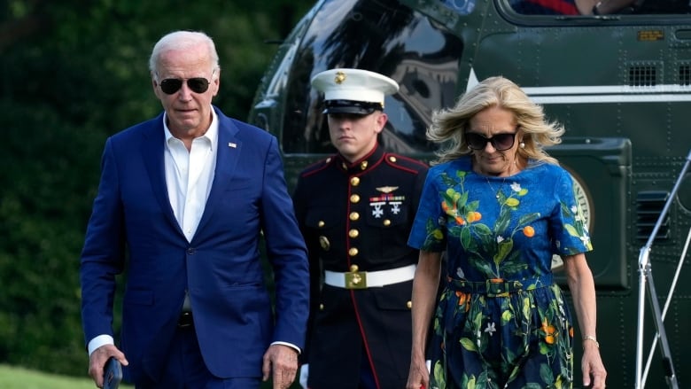 An older man and woman, each wearing sunglasses, and he wearing a suit jacket and her wearing a dress, are shown near a military guard in uniform and a helicopter.