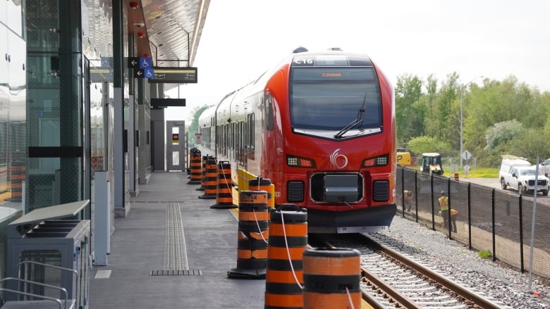 A red and white train stops at a station platform lined with pylons while people in orange vests work in the background