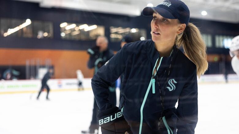 A blonde woman in a ball cap looks at someone while standing on ice.