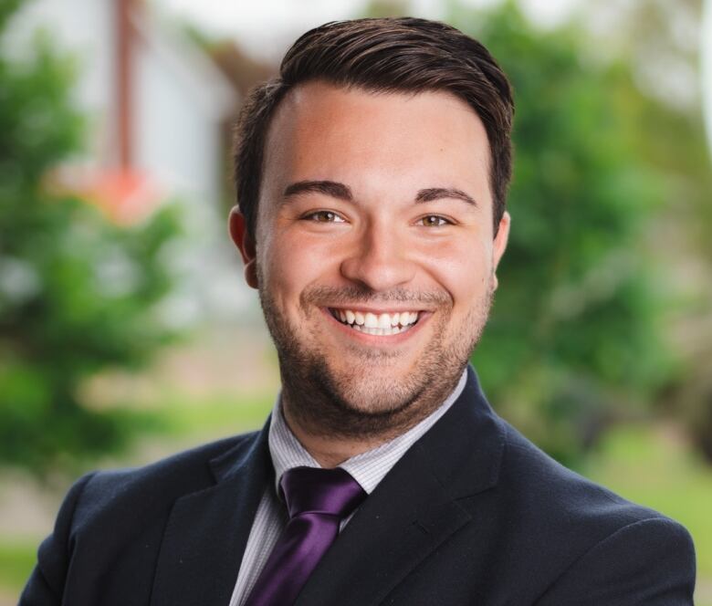 A man with short brown hair, parted on the side and brushed back, and a thin beard and moustache, wearing a black suit, white shirt and purple tie, smiles at the camera for a head and shoulders professional portrait.