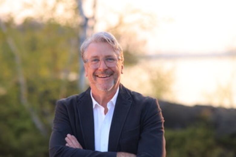 A older white man with grey hair smiling with his arms crossed wearing a black blazer and white dress shirt. He's standing outside where the leaves behind him are yellow.