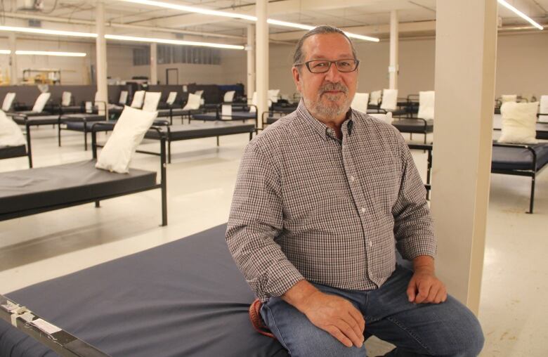 A man in a checkered shirt is seated on a blue bed, in a room full of beds. 