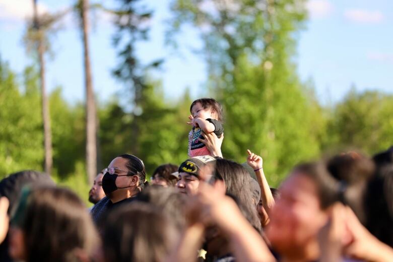 A baby sits on top of shoulders in an outdoor concert crowd. Trees in the background.