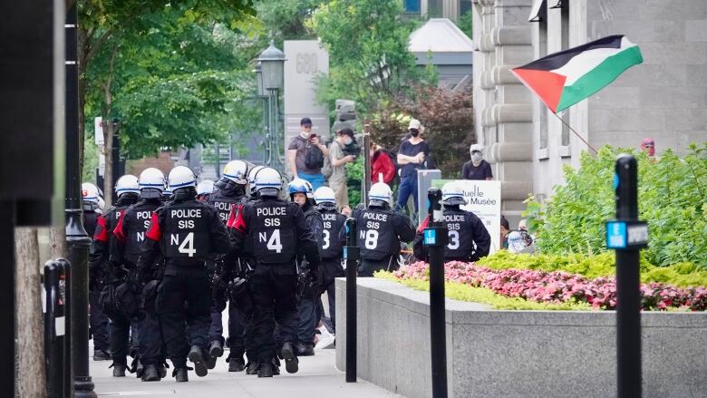 Police in riot gear surround the university as a Palestinian flag waves. 