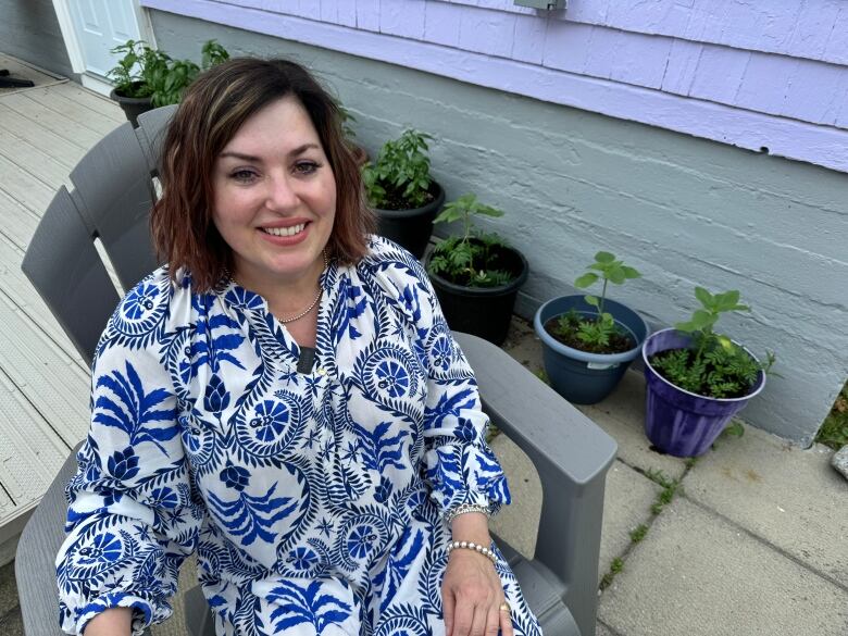 A Caucasian woman in a white and blue dress sits on a patio chair on an outdoor deck. Behind her are potted basil plants. 