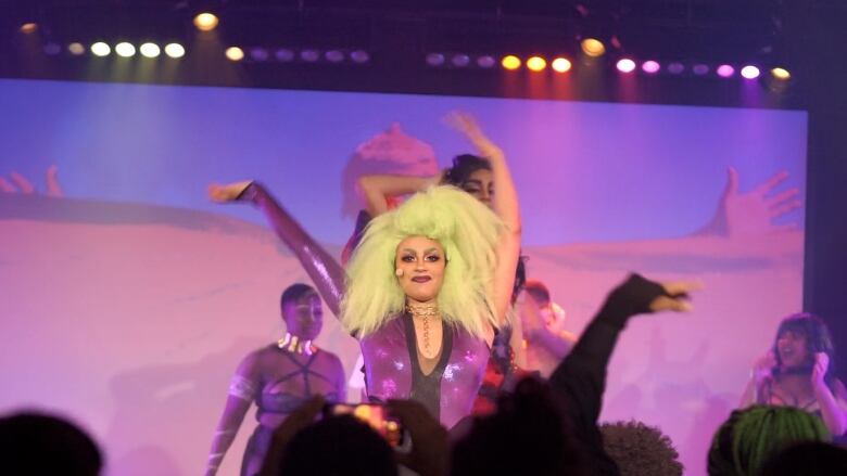 A person with a big green wig and purple shimmery body suit stands on stage with other performers.