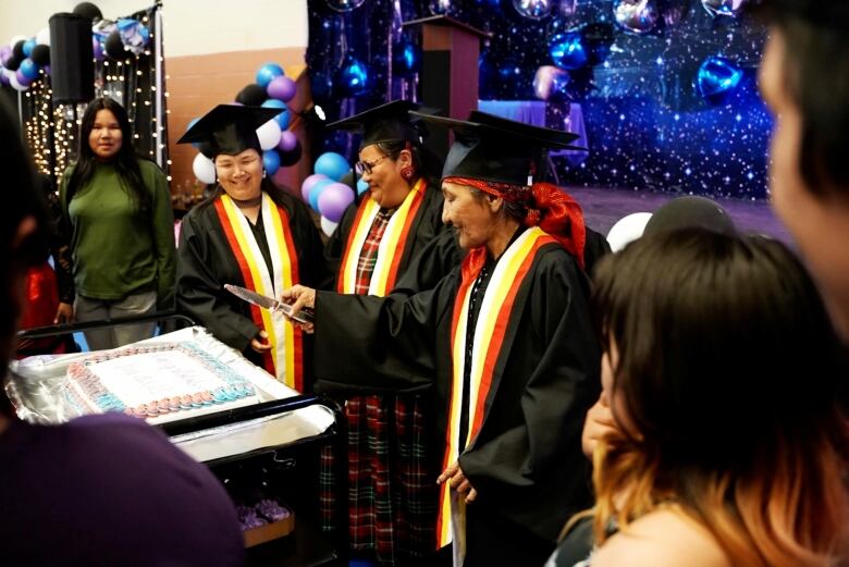 Four women in graduation robes look toward a pink and blue cake that says Congratulations Graduates. 