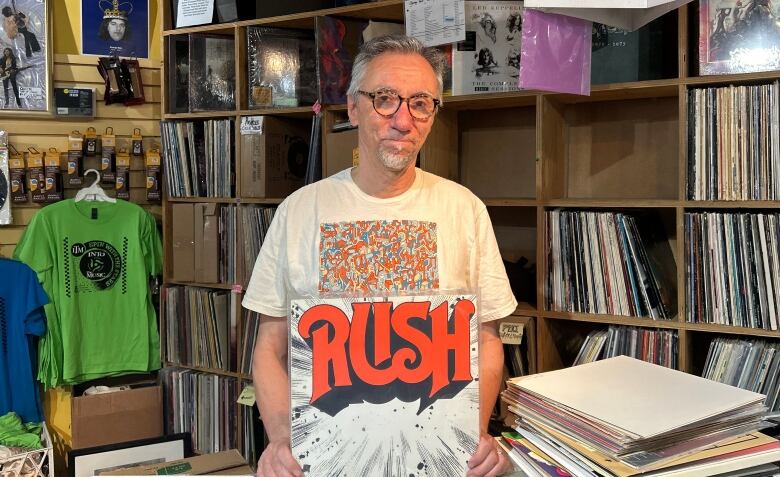 A man with glasses holds a vinyl with his hands in front of shelves stacked with more vinyls.