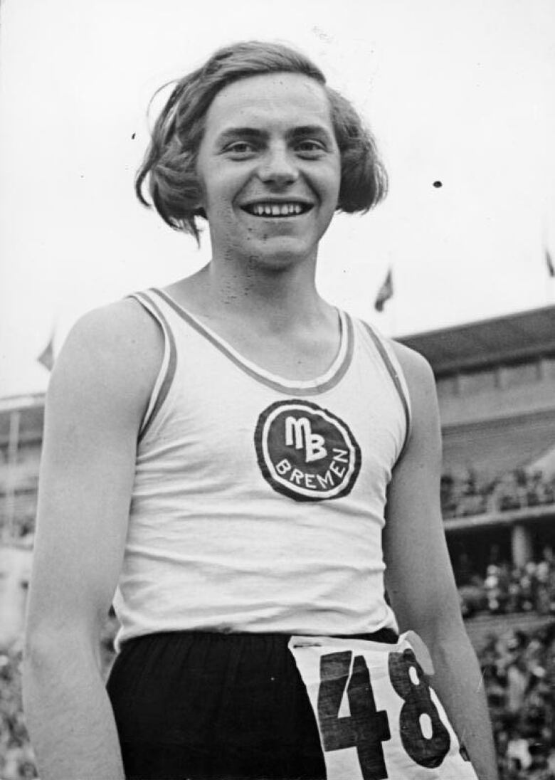 A person in a white tank top and dark shorts with the number 48 smiles while outside at an athletic competition. 