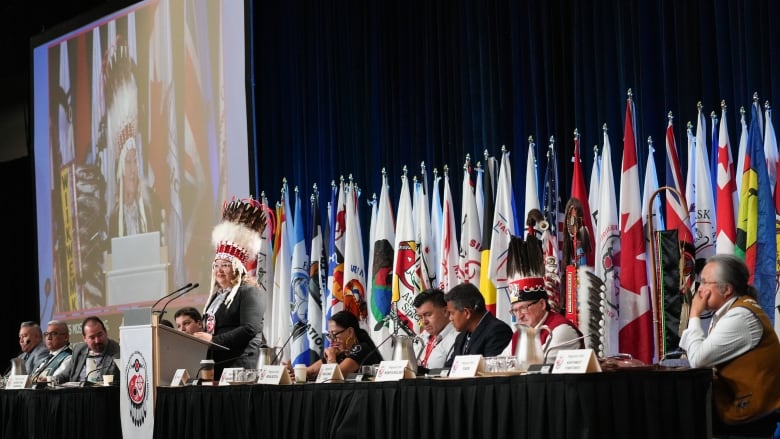 A group of people sit at a black table near a podium. A line of flags are behind them. 