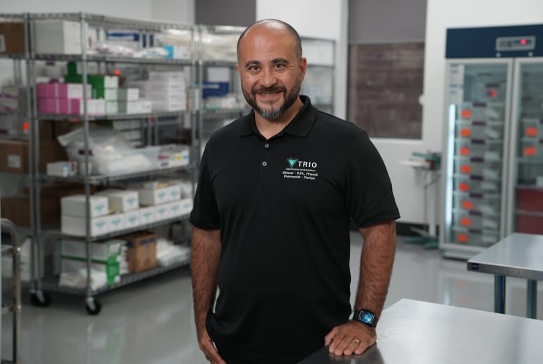 A balding man smiling with a great beard stands wearing a black t-shirt in a workplace with a fridge behind him and shelves to the side. 