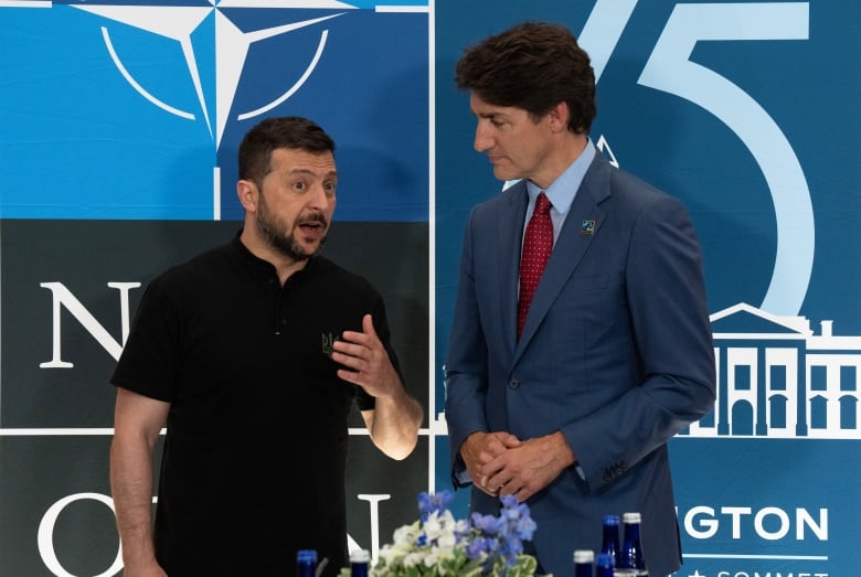 Two men stand in front of a blue backdrop; the one on the left in a black polo shirt and the one on the right in a blue suit with a red tie. 