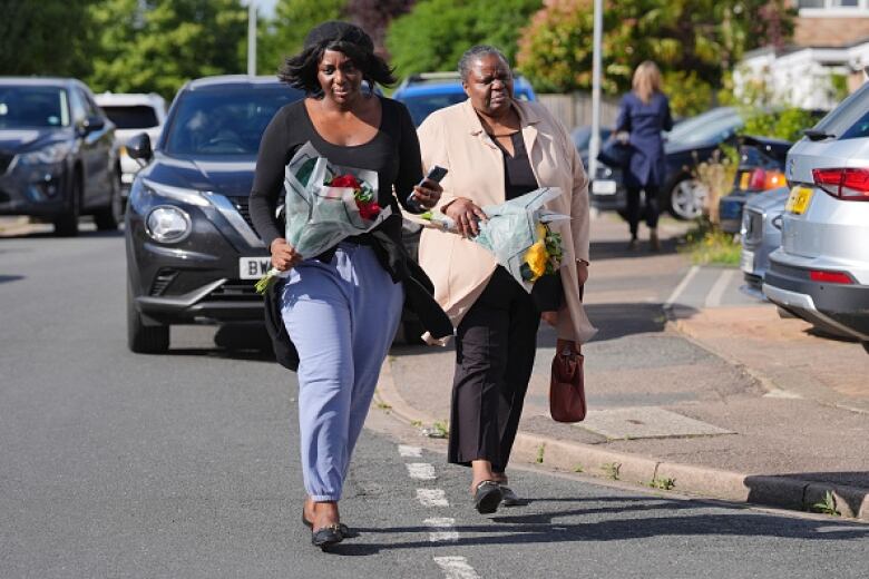 Two women walk on a street carrying bouquets of flowers.