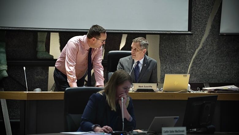 A man in a pink shirt leans forward on a desk where a man in a dark suit is sitting