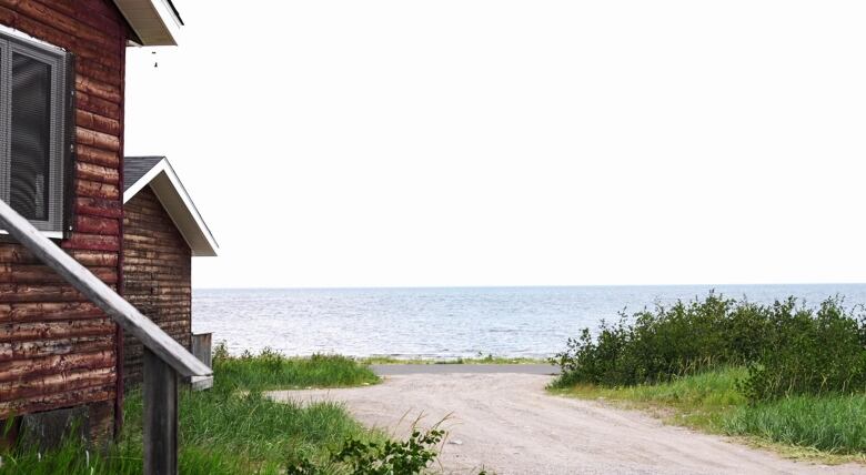 Two brown buildings are in front of a large lake with greenery surrounding it. 