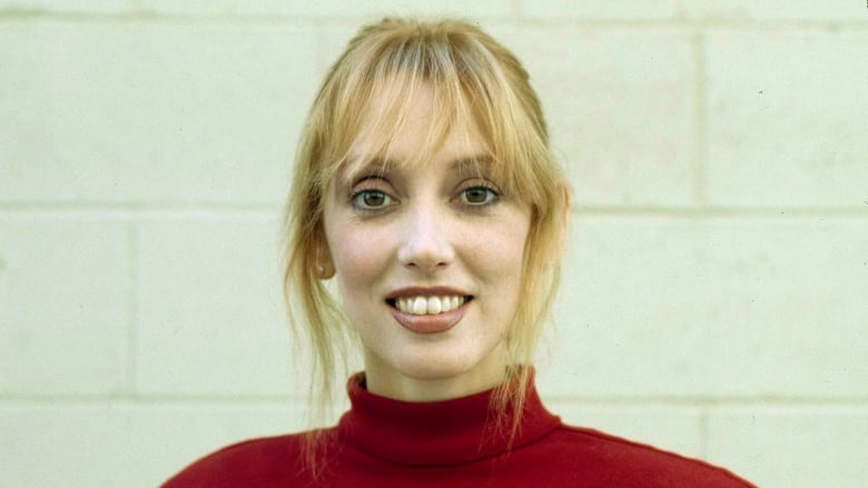 A buck-toothed woman with big eyes in tied-back light-coloured hair stares into the camera.