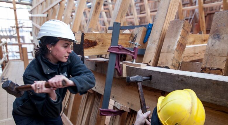 A woman swings a sledgehammer to drive in a spike in a wooden ship's frame.