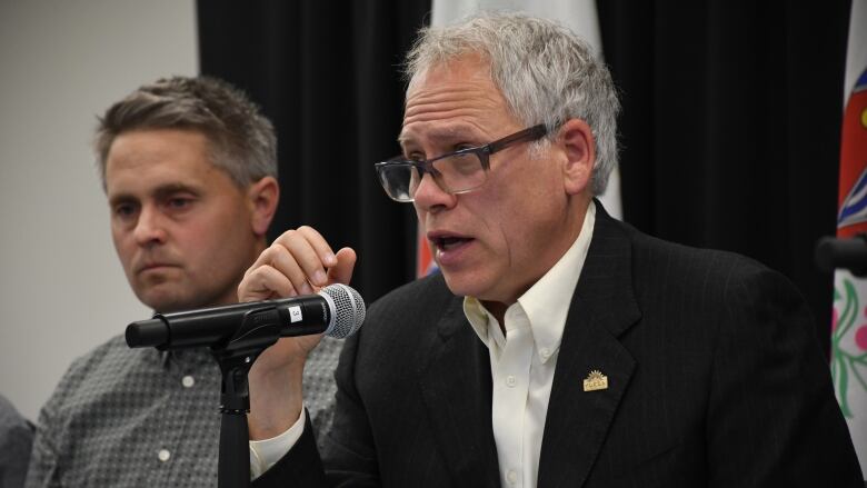 A man speaks into a microphone as another man looks on.