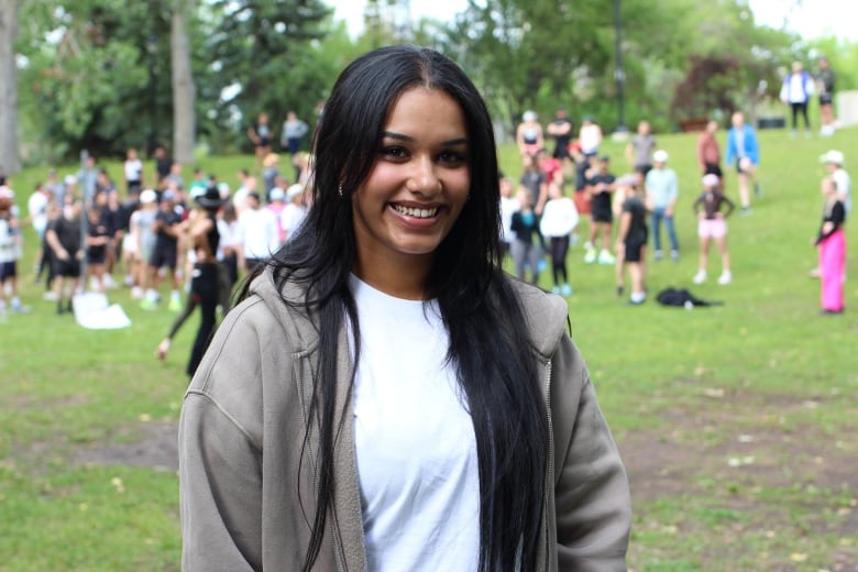 A woman with long black hair, wearing a white t-shirt and hoodie, smiles in a Calgary park in front of a group of runners.