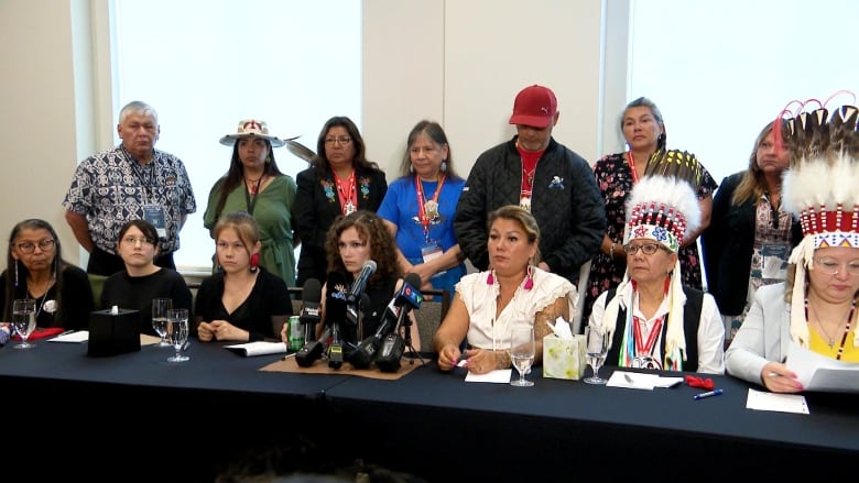 Seven people sit at a table with seven behind standing behind them. 