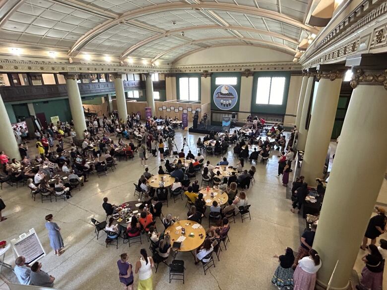 A wide shot shows the interior of a large and crowded room