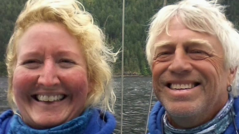 A man and a woman smile for a selfie. Behind them are water and trees.