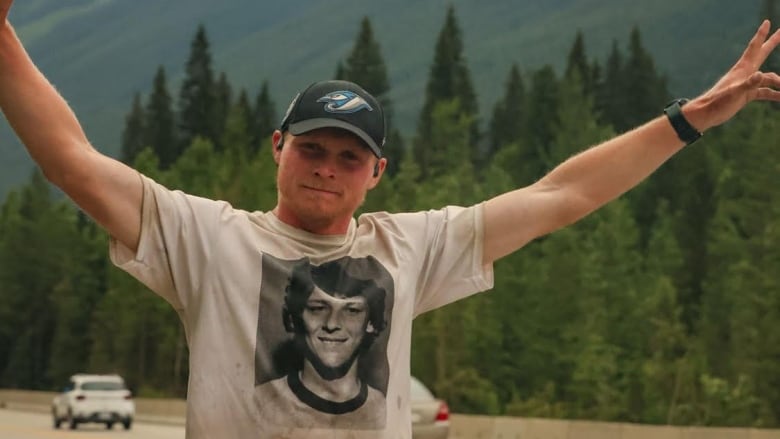 Man stands with his arms stretched out near a roadway, trees and mountains.