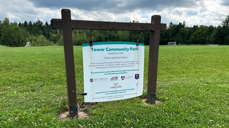 A white and green sign hangs from a brown wooden post in a grassy park surrounded by trees. Two soccer nets can be seen in the distance.