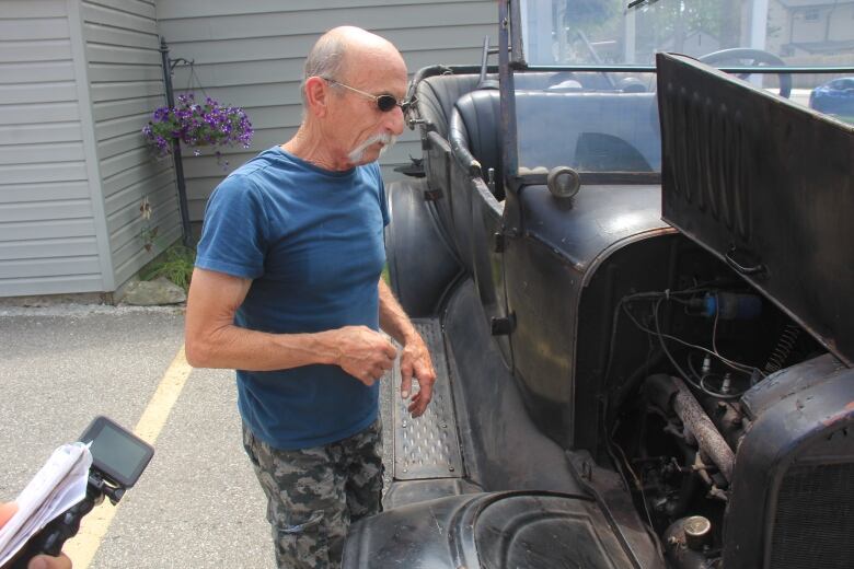 A man with a blue shirt, small sunglasses and a grey mustache inspects under the hood of an old car.