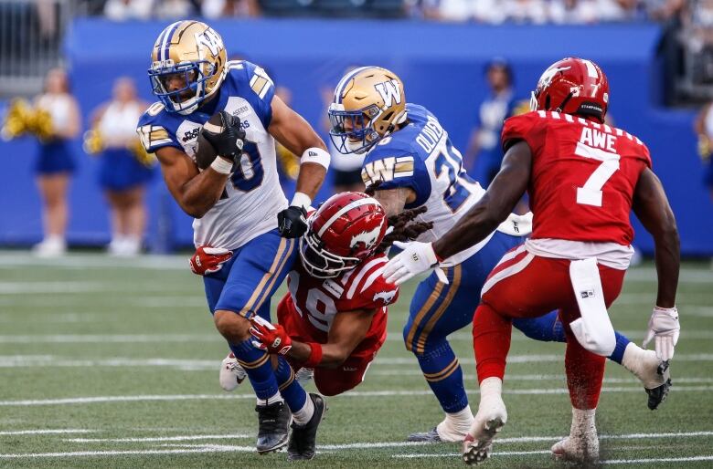 A man wearing a blue and white uniform runs with a football across a stadium while other football players chase after him. 