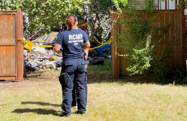 A woman wearing an 'RCMP Fire Investigation Unit' T-shirt speaks to a man in a cordoned-off yard.