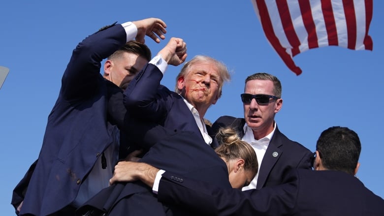 A person with blood on their face raises a fist as they are escorted by security.